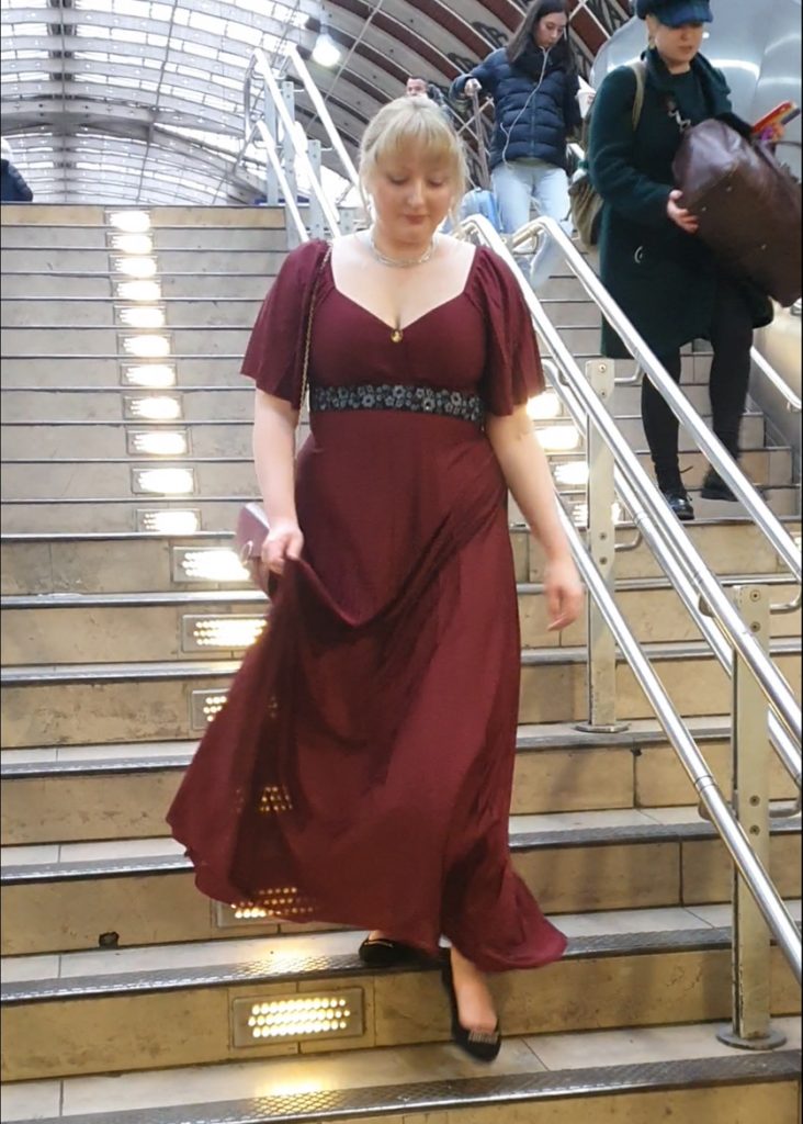 Becky walking down stairs in a station, wearing a long red Regency inspired dress