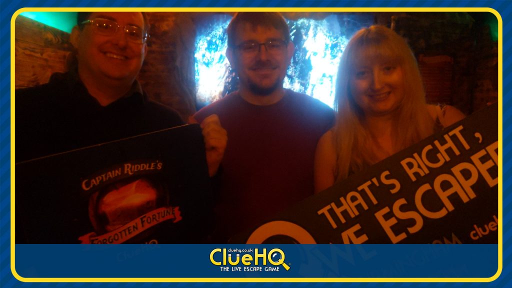 Ed, Becky and Tim smiling in their escape room success photo, Becky is holding a sign reading "That's Right, We Escaped"