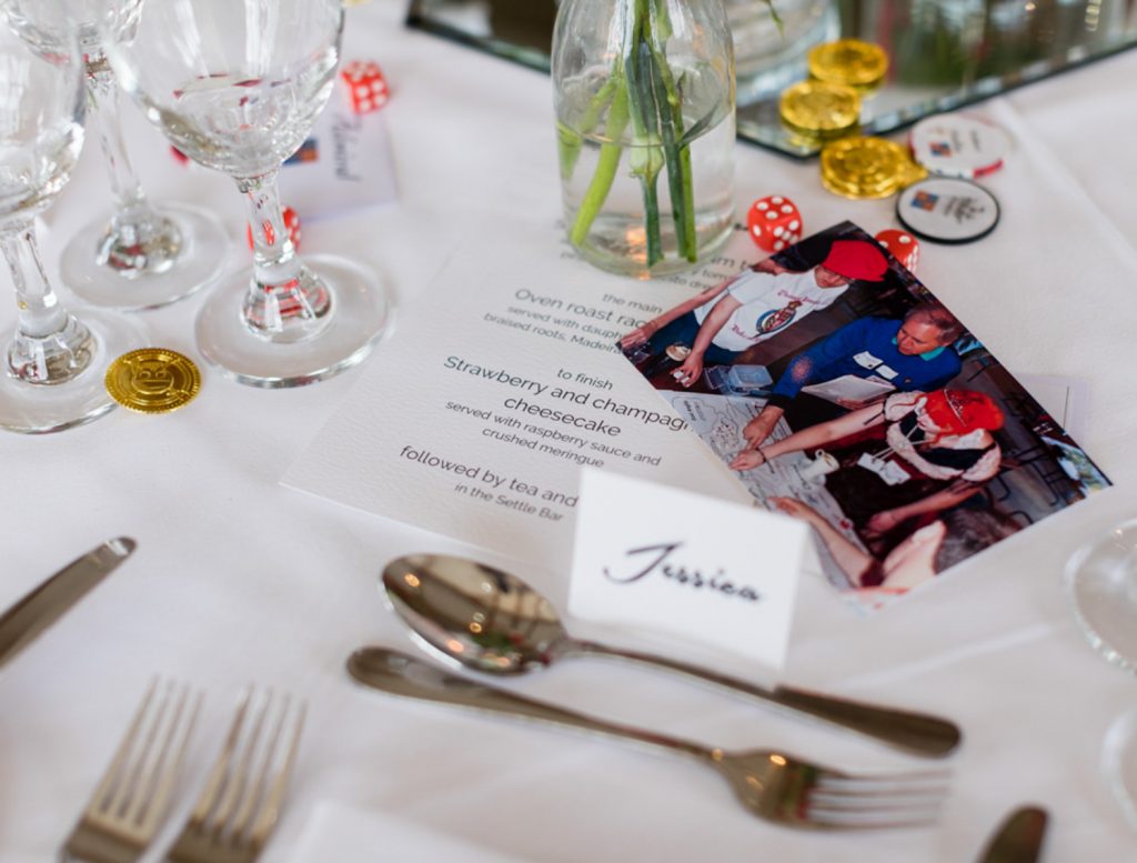 Photo, fake coins and game components from The Spanish Road on a table set with glasses, knives, forks and place name