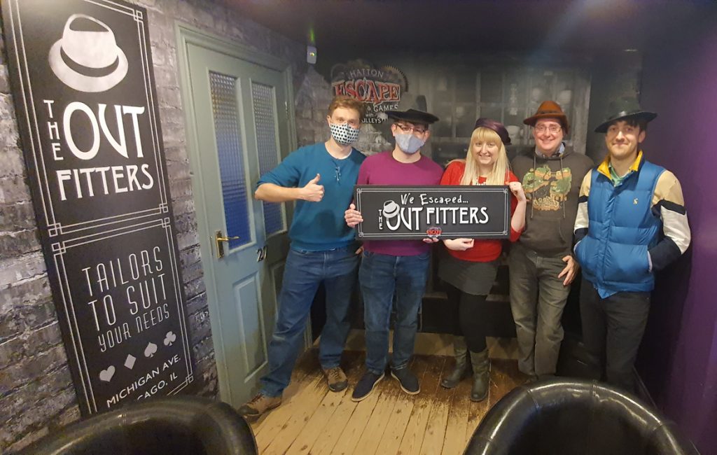Becky and friends holding a sign reading "We escape the Outfitters" in front of the sign to the room
