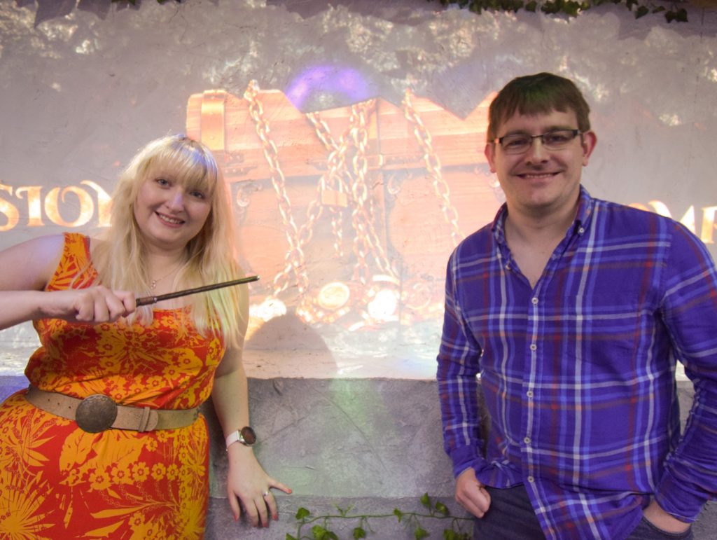 Becky and Tim smiling in front of a screen with a projected treasure chest, Becky is holding a magic wand