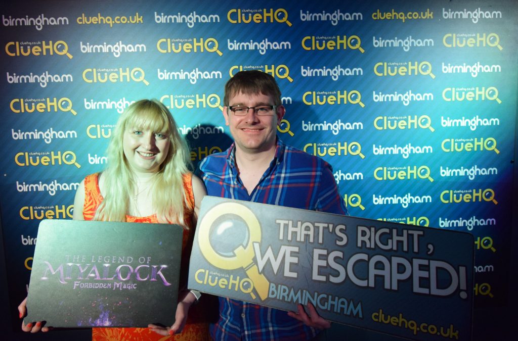 Becky and Tim smiling for their room escape success photo in front of a ClueHQ Birmingham backdrop. Becky is holding a sign reading "The Legend of Miyalock: Forbidden Magic" and Tim's sign reads "That's right, we escaped!"