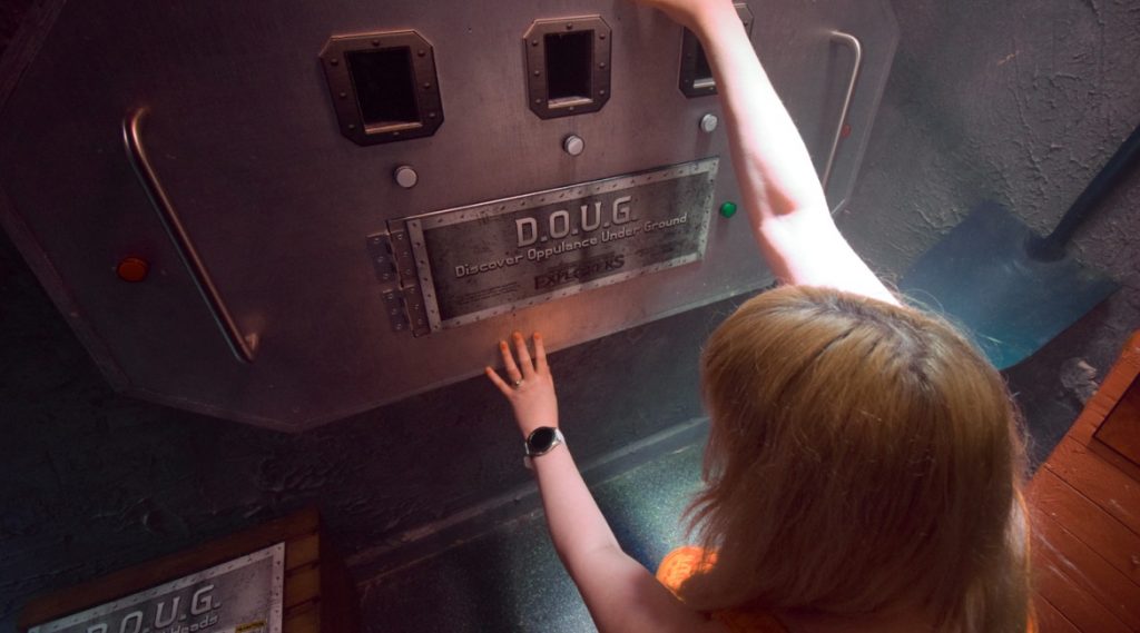 Becky kneeling down and looking at a safe with D.O.U.G. (Discover Oppulance Under Ground) and three holes with buttons