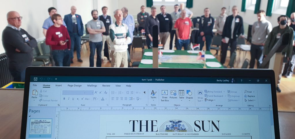 A room full of megagame players, viewed over the top of a laptop screen reading "The Sun"