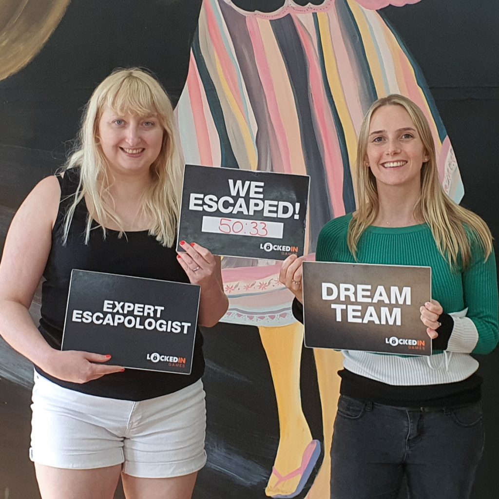Becky and Ellie smile and hold up signs reading "Dream Team", "Expert Escapologist" and "We Escaped: 50:33"