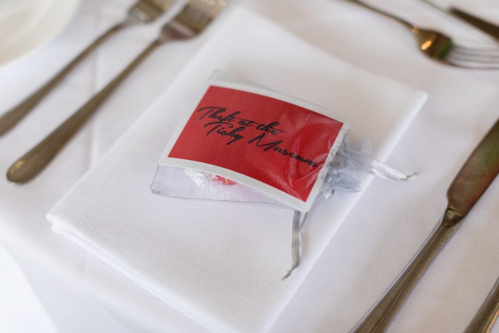 A favour bag on a napkin containing a red card reading "Theft at the Ticky Museum"